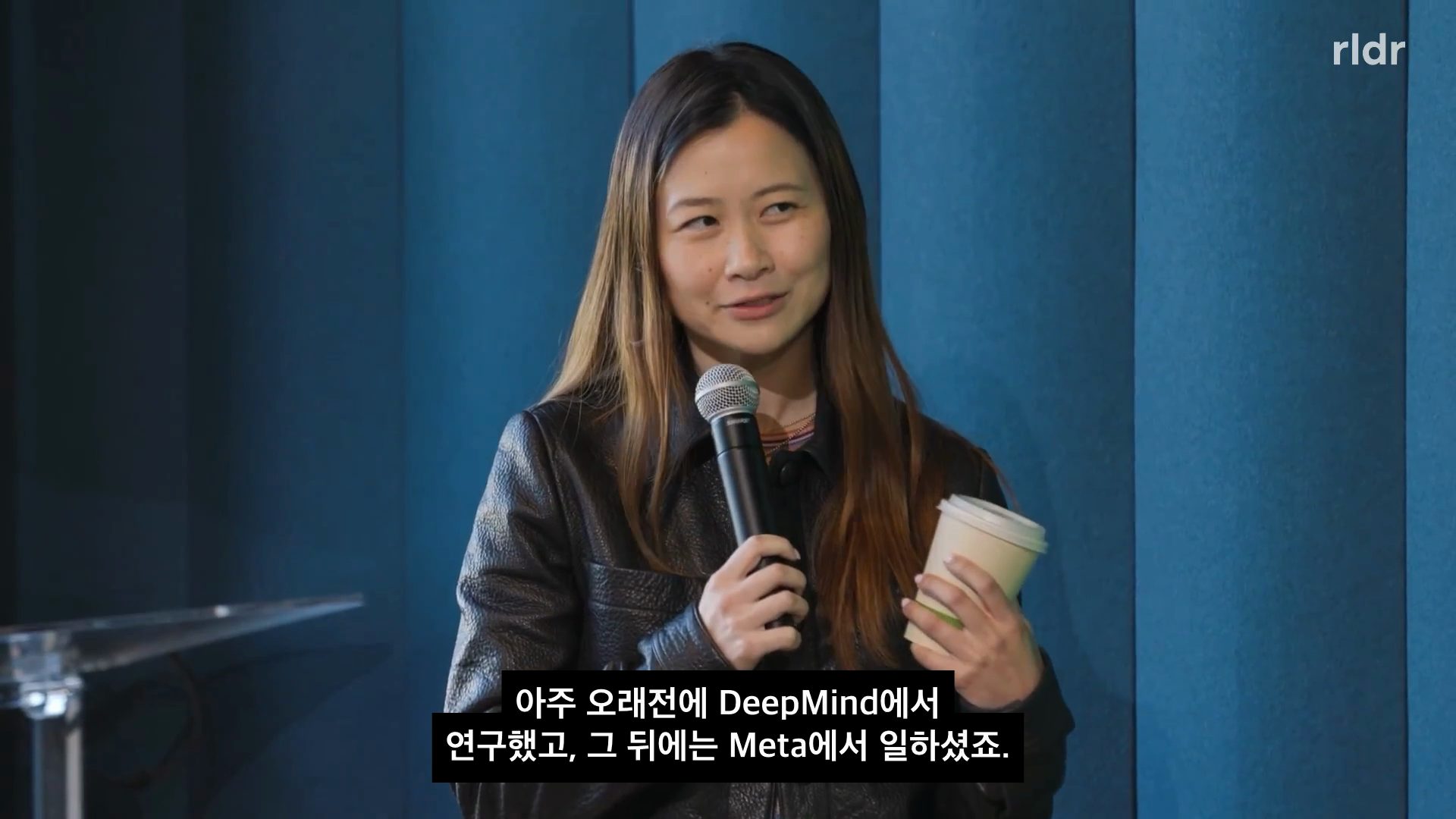 Harvey 발표를 소개하는 진행자가 마이크를 들고 있는 RLDR 영상 장면